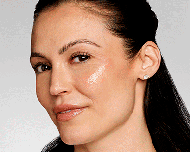 A close-up of a woman with long brown hair with a swipe of moisturizer across her cheekbones, on a gray background.